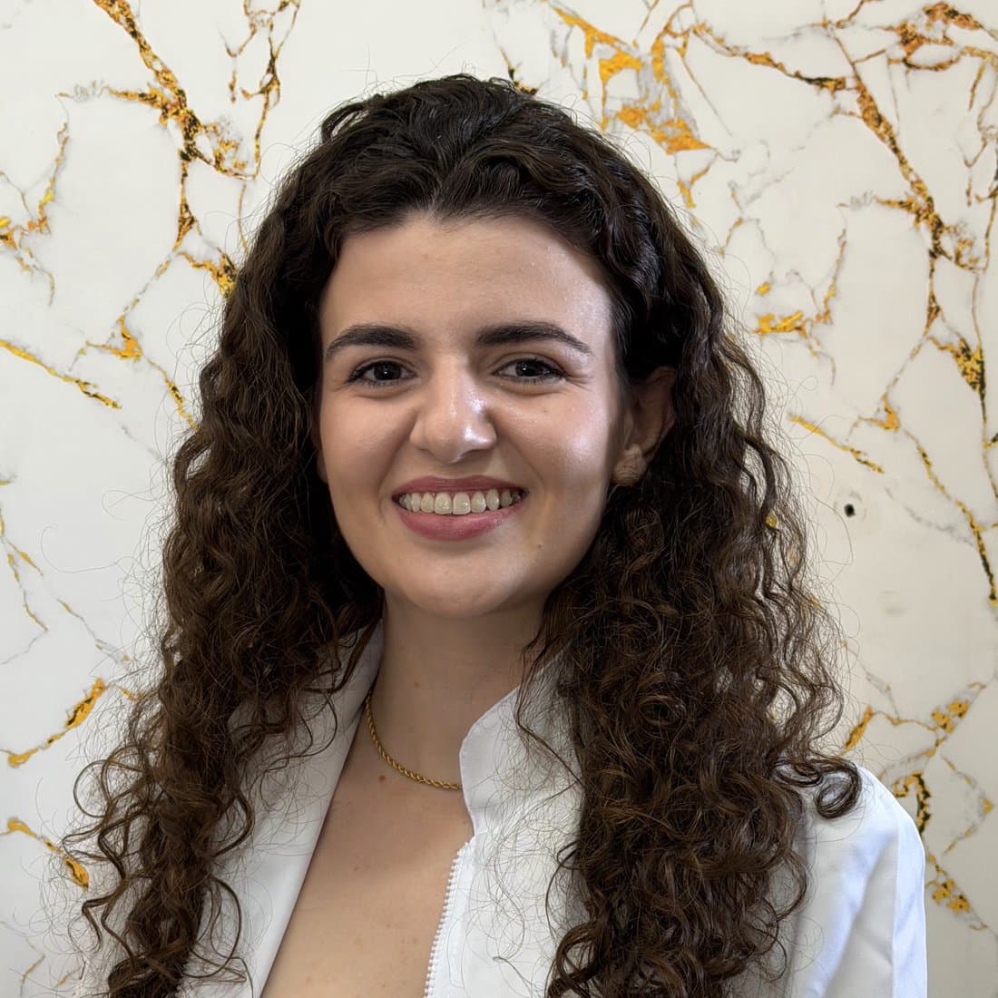 Leiziane Sartti sorrindo, de jaleco branco, em frente a um painel de mármore