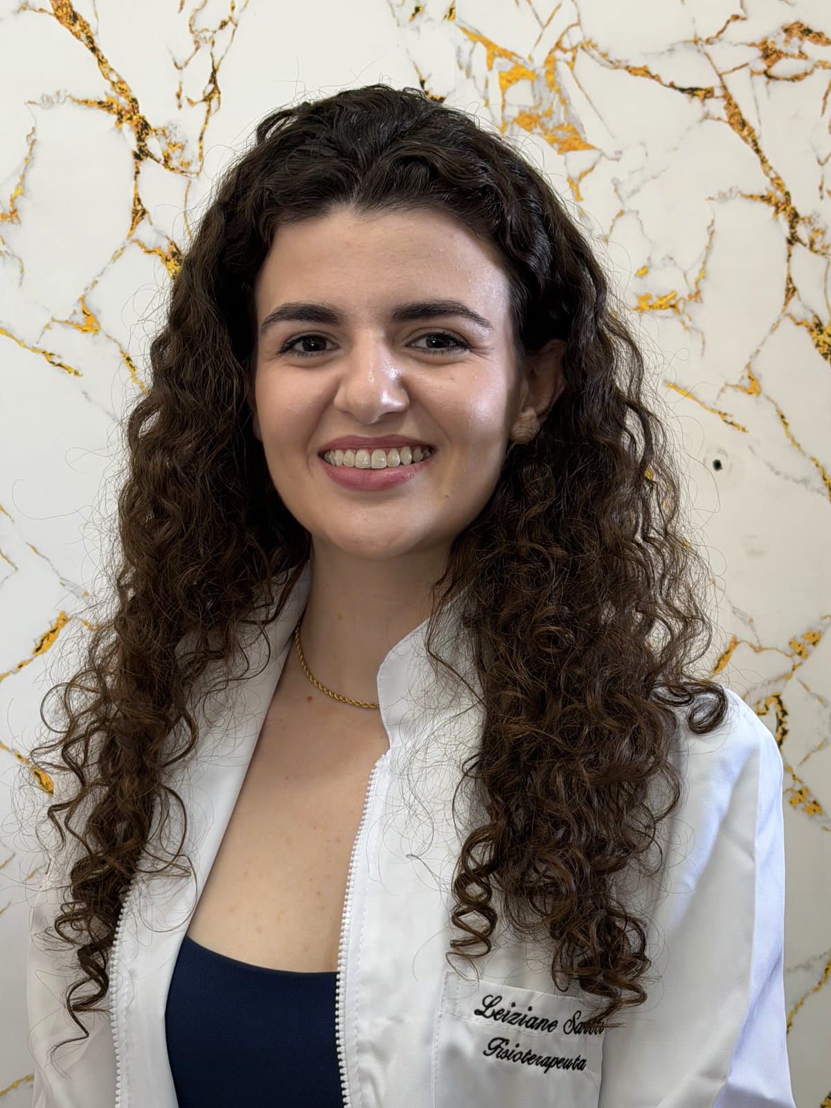 Leiziane Sartti de jaleco branco em seu espaço de atendimento, sorrindo para a câmera